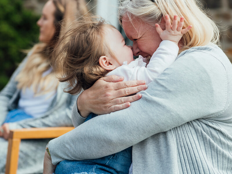 A senior woman embracing her granddaughter and laughing. They are wearing casual clothing and are sitting in a garden at a baby shower.