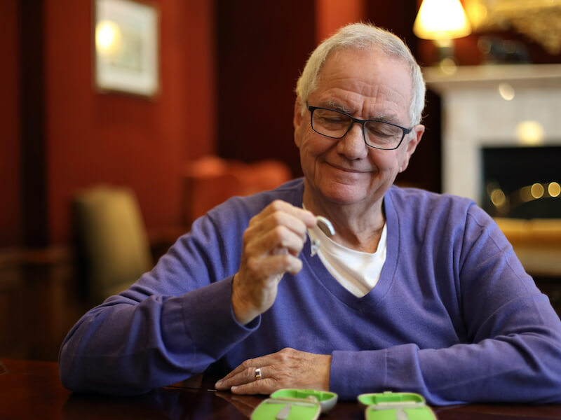 Man wearing purple shirt sitting at a table with his new hearing aids examining them and smiling.